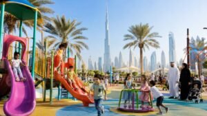 Best playgrounds in Dubai featuring children playing in a modern outdoor playground with colorful climbing structures, slides, splash park, palm trees, and the Dubai skyline in the background.